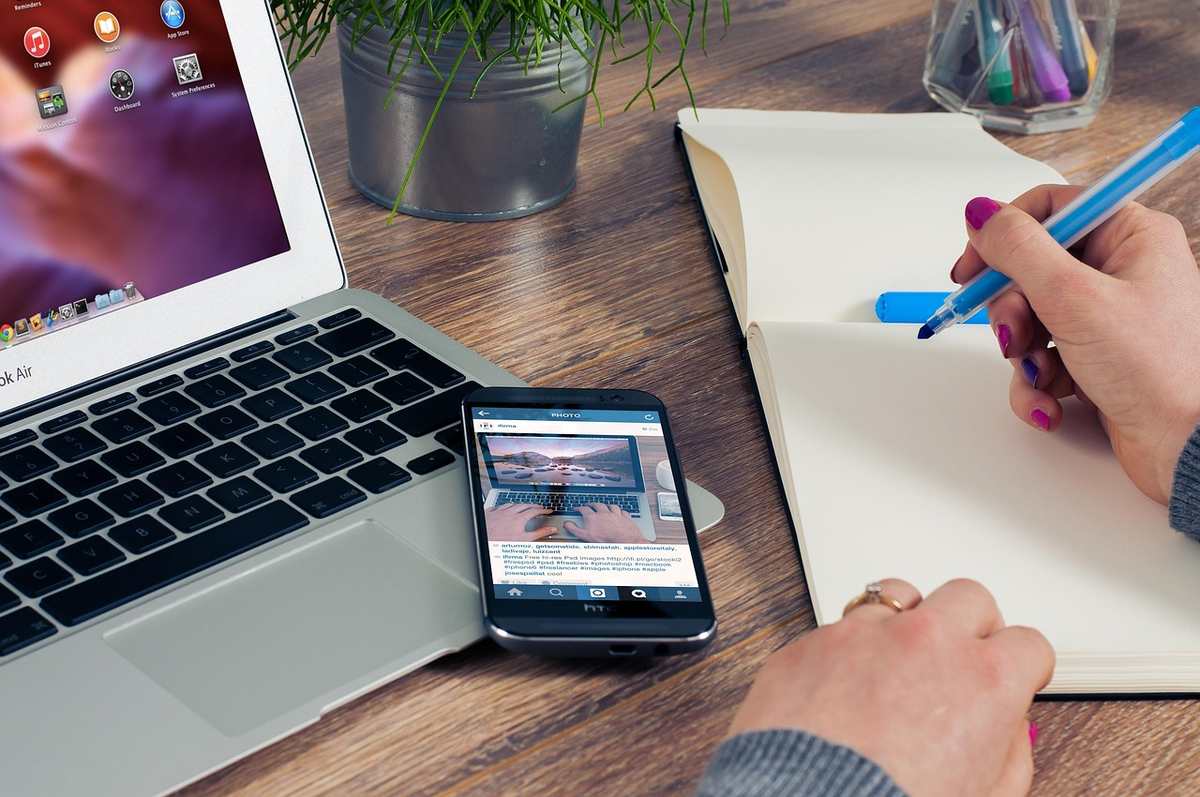 Une personne utilise un stylo pour écrire dans un carnet à côté d'un smartphone et d'un ordinateur portable sur une table en bois
