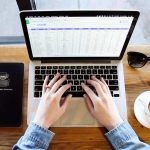 Personnes mains au clavier d'un ordinateur portable sur une table en bois avec un carnet, des lunettes de soleil et une tasse de café