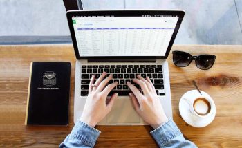 Personnes mains au clavier d'un ordinateur portable sur une table en bois avec un carnet, des lunettes de soleil et une tasse de café